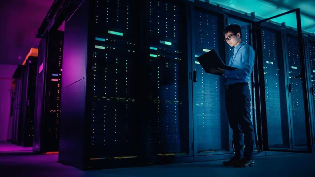 In Data Center: Male IT Technician Running Maintenance Programme on a Laptop, Controls Operational Server Rack Optimal Functioning. Modern High-Tech Operational Super Computer